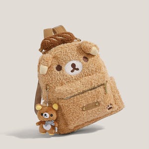 May include: A brown teddy bear-themed backpack with a plush, textured exterior. The backpack features a small front pocket, a bear face design, and a matching bear charm. The straps are brown, and the backpack has a small, brown bread roll on top.