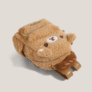 May include: A brown teddy bear backpack with a zipper closure and adjustable straps. The backpack has a plush, furry texture and features a bear face with embroidered details. The straps and accents are a light brown color.
