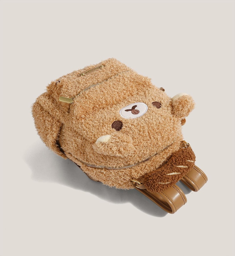 May include: A brown teddy bear backpack with a zipper closure and adjustable straps. The backpack has a plush, furry texture and features a bear face with embroidered details. The straps and accents are a light brown color.
