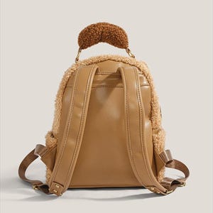 May include: A tan backpack with a fluffy, teddy bear-like texture around the edges and a brown, textured handle. The back of the backpack is smooth tan leather with adjustable straps and a small pocket.