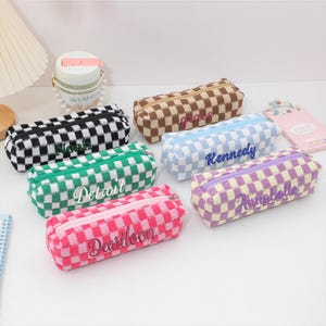 May include: A collection of six rectangular pencil cases featuring a checkered pattern in black, brown, light blue, green, pink, and purple. Each case has a zipper closure and is embroidered with a unique name: Noah, grace, Kennedy, Detroit, Annabelle, and Dearloon.