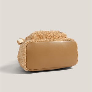 May include: A tan-colored handbag with a teddy bear-like texture on the top half and a smooth, light brown bottom. The bag has a small, rounded handle on the side. The overall design is soft and plush, suggesting a casual or playful style.