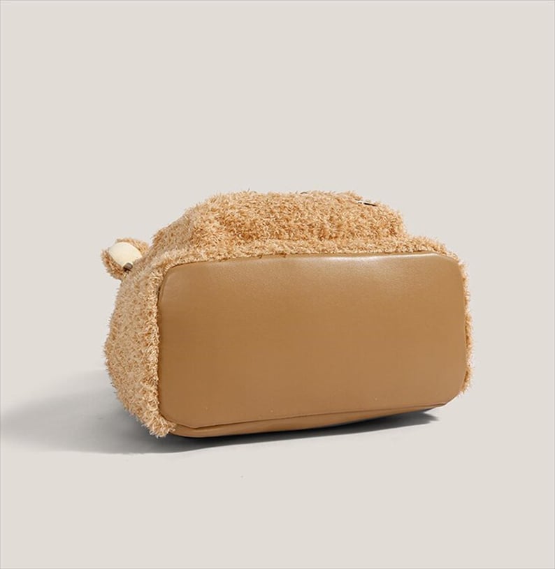 May include: A tan-colored handbag with a teddy bear-like texture on the top half and a smooth, light brown bottom. The bag has a small, rounded handle on the side. The overall design is soft and plush, suggesting a casual or playful style.