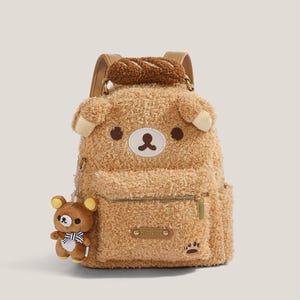 May include: A brown plush backpack shaped like a bear with a small bear charm attached. The backpack has a zipper pocket, paw print detail, and a bread-shaped handle. The bear's face features brown eyes and a white and brown nose.