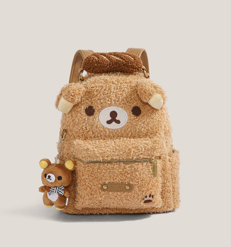 May include: A brown plush backpack shaped like a bear with a small bear charm attached. The backpack has a zipper pocket, paw print detail, and a bread-shaped handle. The bear's face features brown eyes and a white and brown nose.