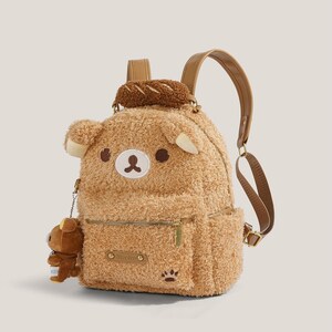 May include: A brown plush backpack shaped like a bear, featuring a small bear charm, a bread-shaped accessory, and a paw print detail. The backpack has a zipper closure and adjustable straps.