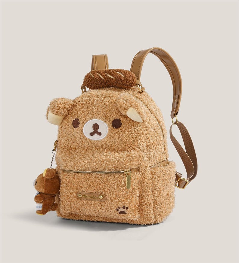 May include: A brown plush backpack shaped like a bear, featuring a small bear charm, a bread-shaped accessory, and a paw print detail. The backpack has a zipper closure and adjustable straps.