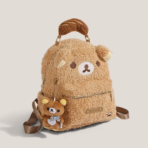May include: A brown plush backpack with a bear face design, featuring a small bear charm. The backpack has a top handle and adjustable straps. The design includes a zipper closure and decorative stitching.