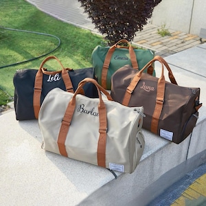 Op de afbeelding: Vijf gepersonaliseerde duffeltassen in verschillende kleuren: zwart, groen, bruin en beige. Elke tas heeft bruine handvatten en riemen, met een naam in sierlijke letters geborduurd. De tassen worden buiten op een betonnen oppervlak getoond.