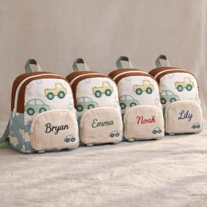 May include: Four personalized children's backpacks. Each backpack features a brown top, a white front with car illustrations, and a light blue side panel with car patterns. The front pocket is embroidered with a name: Bryan, Emma, Noah, and Lily.