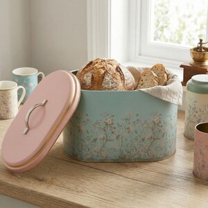 Bread box for kitchen counter showing a large oval farmhouse bread bin filled with fresh sourdough bread loaves and slices on a wooden tabletop, featuring a vintage botanical floral design with a pink lid next to matching kitchen canisters and coffee mugs.