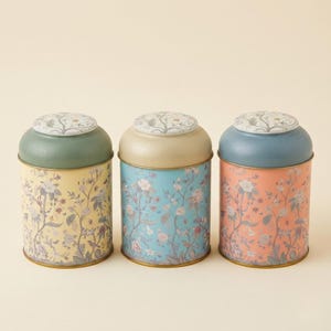 > Spice jar set and vintage kitchen canister set for countertop organization, featuring three decorative metal storage jars with botanical wildflower prints in pastel yellow, light blue, and coral pink colors with porcelain lids, isolated on a clean studio background for farmhouse cottagecore home decor.