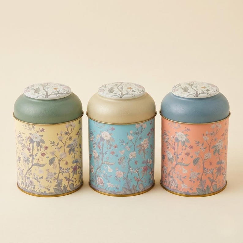 > Spice jar set and vintage kitchen canister set for countertop organization, featuring three decorative metal storage jars with botanical wildflower prints in pastel yellow, light blue, and coral pink colors with porcelain lids, isolated on a clean studio background for farmhouse cottagecore home decor.