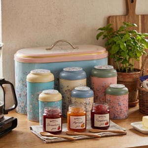 Coffee station decor kitchen canister set on a breakfast table, featuring a vintage blue bread box, floral metal canisters with ceramic lids, and small jam jars with wooden spoons on a countertop next to a coffee maker and basil plant for cottagecore farmhouse kitchen style. (303 karakter)