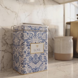 May include: A rectangular metal storage canister featuring a blue and white damask pattern. The front displays a rectangular label with a gold border, a small bee icon, and the word LOVE printed in black text. It has a clear lid with a metal latch.