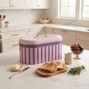 May include: An oval metal bread box with a light pink lid featuring a gold handle and a sunburst design. The body of the container has vertical purple and light pink stripes. The box is placed on a white countertop with toast, butter, and herbs.