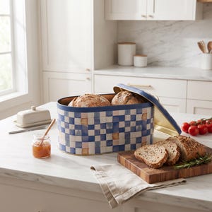 Può includere: Un portapane ovale in metallo con un motivo a scacchi blu, bianco e beige. Il coperchio è parzialmente aperto e rivela due pagnotte all'interno. Un tagliere in legno con pane affettato, un vasetto di marmellata e un burriera sono vicini.