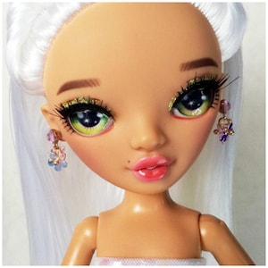 Könnte beinhalten: Das Gesicht einer Puppe mit grünen und gelben Augen, langen Wimpern und rosa Lippen. Die Puppe hat weißes Haar und trägt kleine, baumelnde Ohrringe mit rosa und blauen blütenförmigen Perlen. Die Haut der Puppe ist hellbraun.