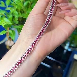 Op de afbeelding: Een zilveren armband met een rij vierkante, dieprode edelstenen, afgewisseld met kleine, heldere stenen. De armband wordt getoond tegen een achtergrond van groen gebladerte.