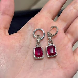 Könnte beinhalten: Ein Paar silberne Ohrringe mit rechteckigen, tiefroten Edelsteinen. Jeder Edelstein ist von einem Kranz aus kleinen, klaren Steinen umgeben. Die Ohrringe werden von einer Hand gehalten.