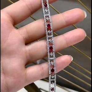 May include: A silver bracelet featuring alternating oval-cut red gemstones and small, round, clear gemstones. The bracelet has a delicate, openwork design with a rectangular shape and is held in a hand.