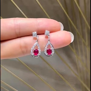 May include: A pair of elegant drop earrings featuring a teardrop-shaped ruby gemstone, surrounded by a halo of small, sparkling white stones. The earrings have a square-shaped top with additional small stones, all set in a silver-colored metal.