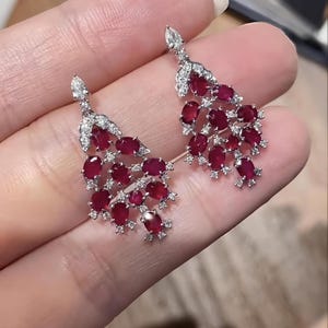 Op de afbeelding: Een paar elegante oorbellen met clusters van ovaal geslepen, dieprode edelstenen, geaccentueerd met kleine, sprankelende witte stenen. De oorbellen hebben een trapsgewijs ontwerp, met een peervormige steen aan de bovenkant. De zetting lijkt zilver of witgoud te zijn.