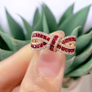May include: A bow-shaped ring with a silver band, adorned with rows of small, vibrant red gemstones and interspersed with clear, sparkling stones. The ring's design includes gold-colored accents, creating a luxurious and eye-catching piece of jewelry.