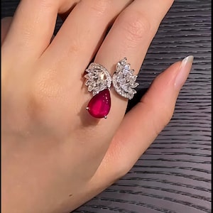 Könnte beinhalten: Silberner Ring mit einem tropfenförmigen Rubin und einem floralen Design. Der Ring hat eine Ansammlung kleiner, klarer Steine um den Rubin und am Ringband.