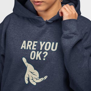May include: Navy blue hoodie with the text "ARE YOU OK?" in white, along with a graphic of a hand. The hoodie has a drawstring hood and a kangaroo pocket. The model is wearing silver hoop earrings.