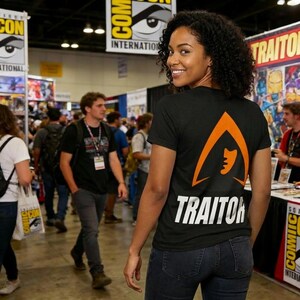 May include: Black t-shirt with an orange graphic and the word "TRAITOR" in white. The shirt is worn by a person at a convention with "COMIC" signage in the background.