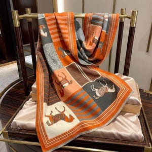 Puede incluir: Bufanda naranja, gris y marrón con un estampado de caballos. La bufanda está colocada sobre un estante dorado. El diseño tiene un estilo patchwork con ilustraciones de caballos en varias poses. El borde es naranja con un ribete color crema.