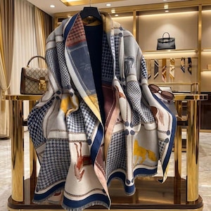 Puede incluir: Una bufanda grande y estampada con una variedad de diseños, que incluyen un patrón de pata de gallo, ilustraciones de caballos y formas geométricas. La bufanda presenta una paleta de colores de azul, blanco, marrón y amarillo. La bufanda está drapeada sobre una prenda de color oscuro.