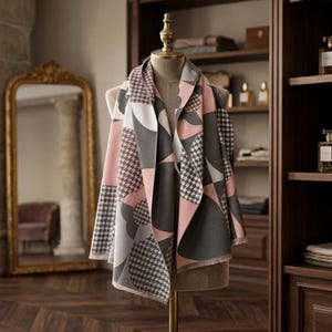 Peut inclure: Une écharpe à motifs dans les tons rose, gris, blanc et noir est drapée sur un mannequin. L'écharpe présente un motif géométrique avec des motifs pied-de-poule et circulaires. L'écharpe est exposée dans une pièce avec un grand miroir et des meubles en bois.