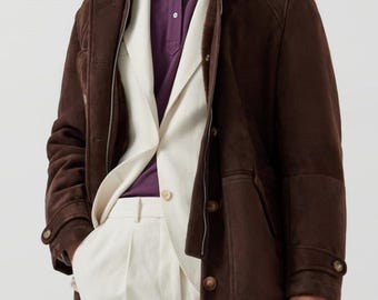 Könnte beinhalten: Eine dunkelbraune Wildlederjacke mit Shearling-Kragen und Knopfverschlüssen. Darunter ein cremefarbener Blazer und eine Hose sowie ein lila Poloshirt. Das Outfit ist ein klassisches, stilvolles Ensemble.