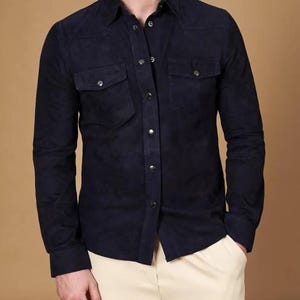 May include: A navy blue suede shirt with long sleeves, two chest pockets, and snap closures. The shirt has a classic collar and is buttoned up. The model is wearing the shirt with cream-colored pants.