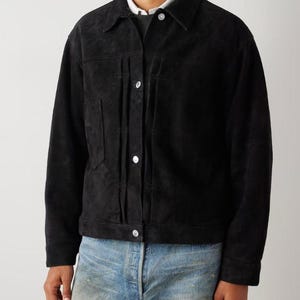 May include: A black suede jacket with a button-up front, two chest pockets, and a collared neckline. The jacket is worn over a light-colored shirt and a gray sweater. The model is wearing blue jeans.