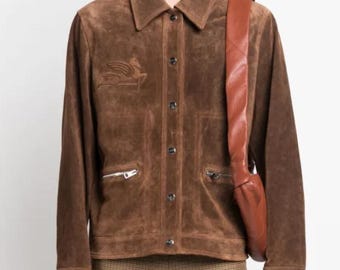 May include: A brown suede jacket with a button-up front, two zipped pockets, and a collared neckline. The jacket features an embroidered design on the left chest and is paired with brown trousers and a brown leather shoulder bag.