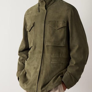 Puede incluir: Chaqueta de ante verde oliva con cuello alzado, dos bolsillos en el pecho y dos bolsillos inferiores. La chaqueta tiene cierre de botones y mangas largas. El modelo lleva pantalones beis.