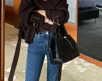 May include: A dark brown jacket with a belt and silver buckles, worn with blue jeans and brown boots. A black quilted handbag is held. The outfit is a fashionable ensemble.