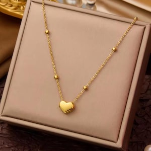May include: A gold-toned necklace featuring a delicate chain with evenly spaced small round beads and a single smooth heart-shaped pendant at the centre. The metal has a polished finish that reflects light, creating a bright and uniform appearance.