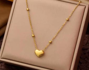 May include: A gold-toned necklace featuring a delicate chain with evenly spaced small round beads and a single smooth heart-shaped pendant at the centre. The metal has a polished finish that reflects light, creating a bright and uniform appearance.