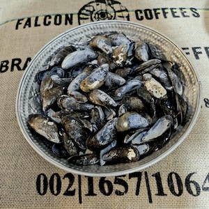 May include: A silver bowl, intricately decorated, overflowing with dark blue mussels. The bowl rests on a hessian sack, printed with "FALCON COFFEES" and numbers. The mussels display varied textures and colours.