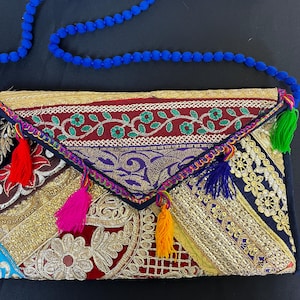 May include: An envelope-style clutch bag featuring a patchwork of embroidered fabrics in red, gold, blue, and black. The flap is adorned with colourful tassels in red, pink, yellow, and blue, and the bag includes a long blue beaded shoulder strap.