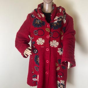 May include: A red, hooded coat with a textured fabric and floral embroidery. The coat features a button-up front and long sleeves. The floral designs include blue, cream, and brown colors. The coat is displayed on a mannequin.
