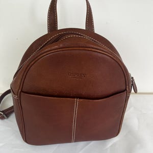May include: A brown leather backpack with a rounded top, a top handle, and a front pocket. The bag features white contrast stitching throughout and the text OSPREY LONDON embossed on the front. It includes a zippered main compartment and side straps.