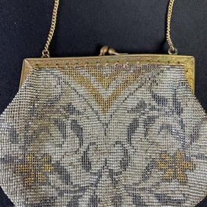 May include: A vintage beaded handbag with a gold-coloured frame and chain handle. The purse displays a detailed pattern in silver, gold, and grey beads. The clasp is a small, decorative metal closure.