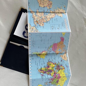 Könnte beinhalten: Eine gefaltete Weltkarte mit detaillierten geografischen Informationen, einschließlich Ländergrenzen, Ozeanen und Städten. Die Karte ist teilweise geöffnet und zeigt ein blaues Lederportemonnaie. Die Karte ist in englischer Sprache.