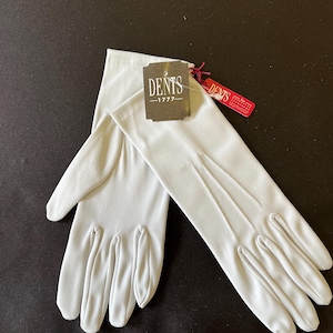Könnte beinhalten: Ein Paar hellgraue Handschuhe mit dem Markenetikett "DENTS -1777-". Die Handschuhe bestehen aus einem glatten Material und haben drei dekorative Linien auf dem Handrücken. Die Handschuhe liegen auf einer schwarzen Oberfläche.