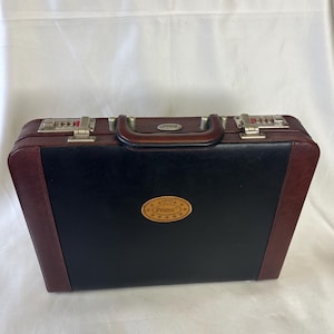 May include: A black and burgundy briefcase with a brown handle and silver latches. The briefcase has a combination lock on each side and a small, oval emblem with the word "Prima" on the front.